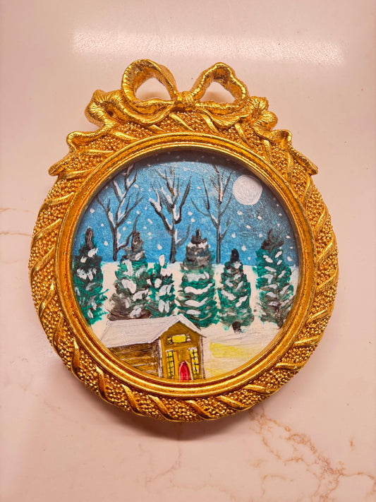 Handmade mini acrylic painting of a snowy winter cabin with pine trees and a full moon in a round gold ornament frame.