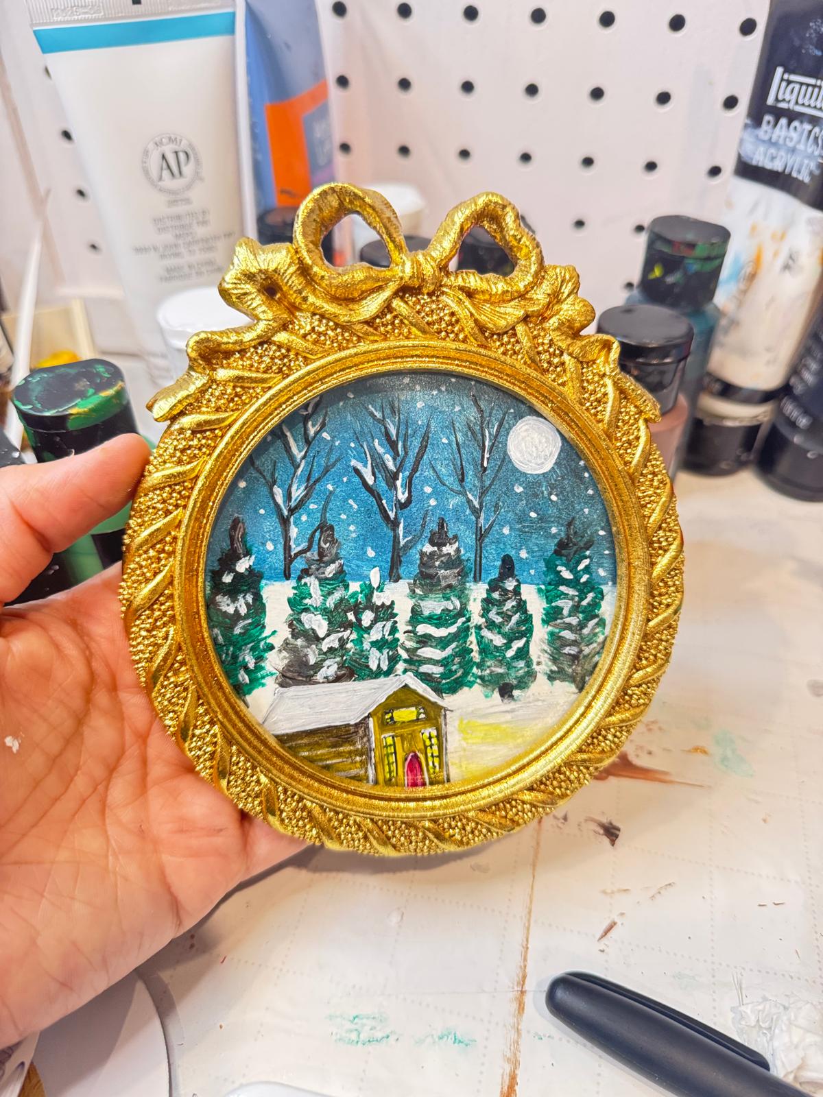 Handmade mini acrylic painting of a snowy winter cabin with pine trees and a full moon in a round gold ornament frame.