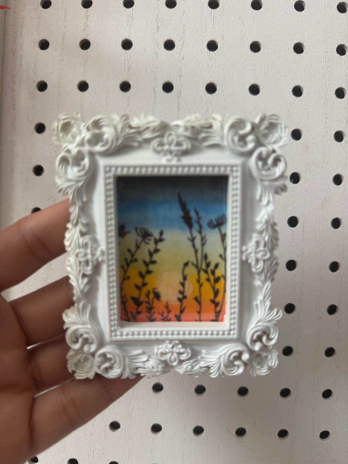 Decorative white frame with a small painting of a sunset scene held against a pegboard background.