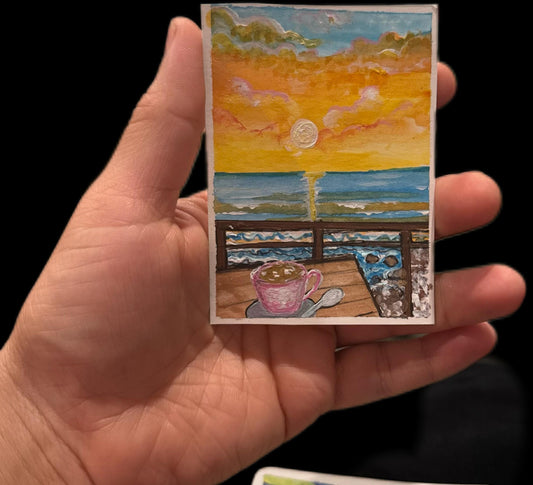 Hand-painted ACEO card of ocean sunset with waves and coffee cup on a deck.