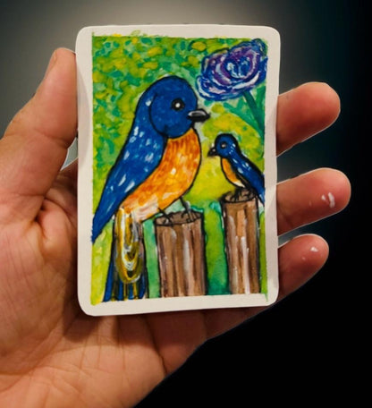 Hand-painted ACEO mini art card of a vibrant blue and orange bird with a smaller bird perched on wooden posts, green floral background.
