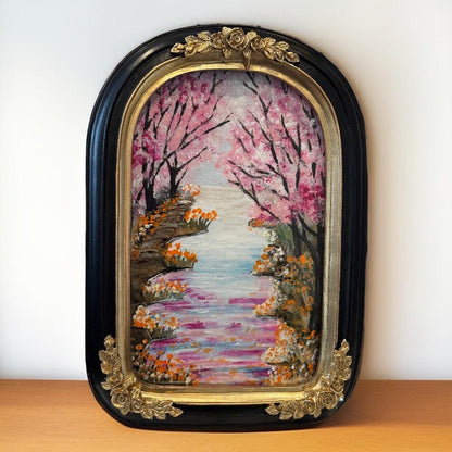 Framed painting “Blossom Pathway” by Faiza Atif – a mixed media artwork of a riverside pathway with pink blossom trees and vibrant flowers in a black and gold ornate frame.