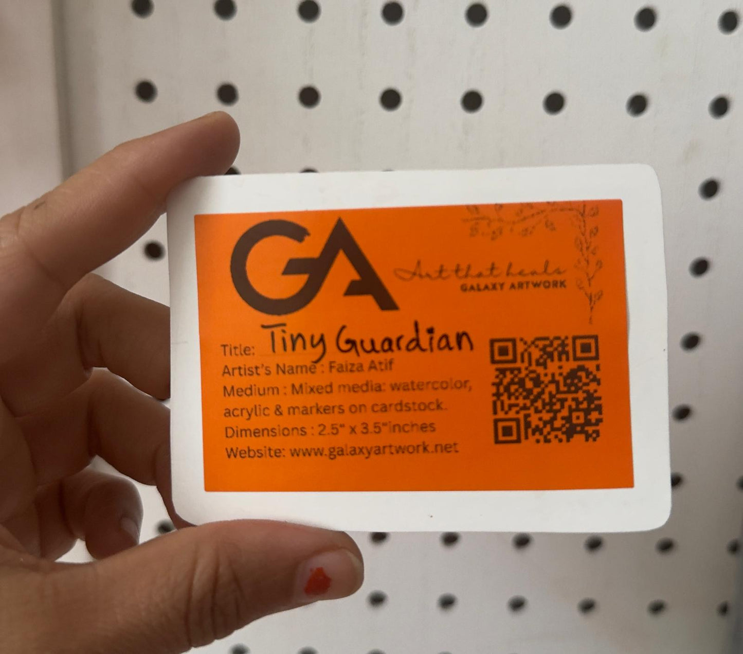 Hand holding a card with 'GA' branding and a QR code against a pegboard background