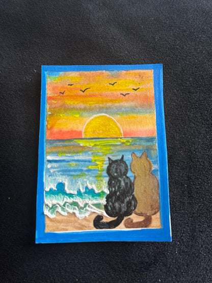 “Two cats sitting on the beach watching a colorful sunset over the ocean with waves and birds in the sky.