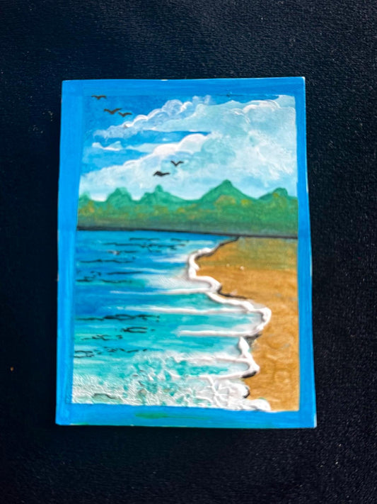 Ocean waves rolling onto sandy beach with distant green mountains and birds flying under a bright blue sky.