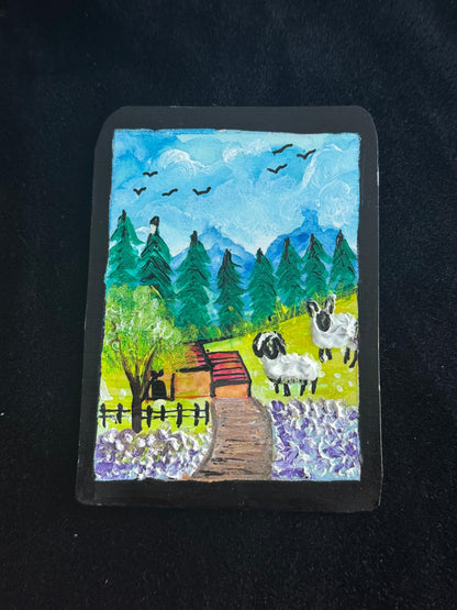 Handmade card with a landscape design featuring sheep, trees, and a cabin on a black background.