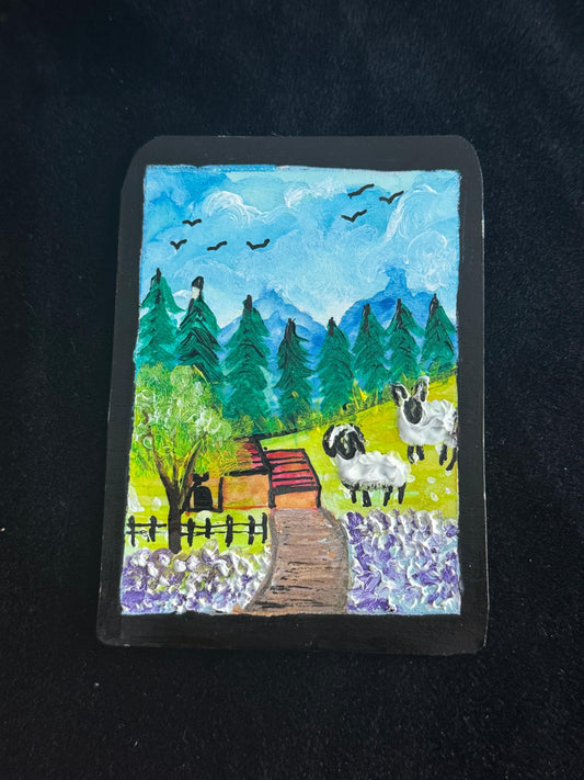 Handmade card with a landscape design featuring sheep, trees, and a cabin on a black background.