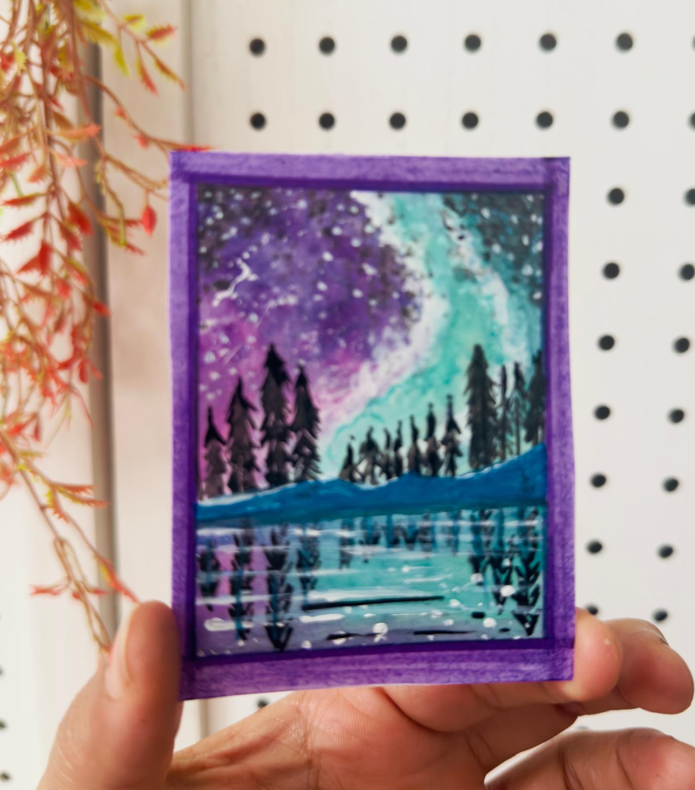 Hand-painted ACEO card of northern lights over lake with pine trees and purple sky.