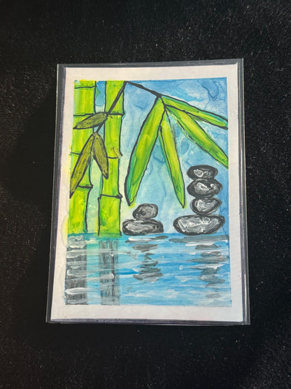 Small hand-painted artwork of bamboo leaves over calm blue water with stacked balancing stones.