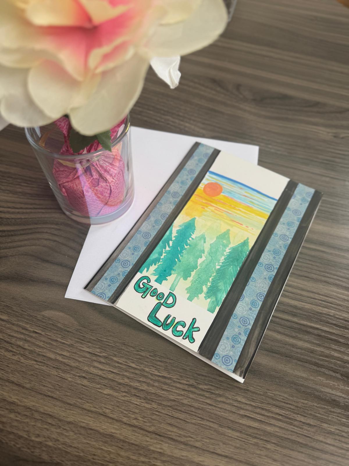 Hand-painted greeting card with pine trees, sunset, and ‘Good Luck’ text, blank inside with envelope