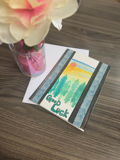 Hand-painted greeting card with pine trees, sunset, and ‘Good Luck’ text, blank inside with envelope