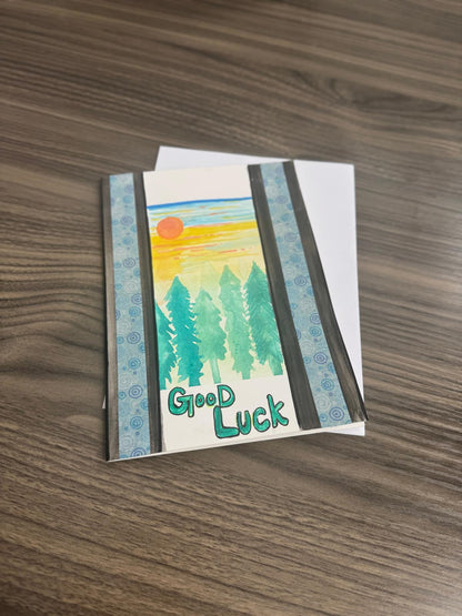 Hand-painted greeting card with pine trees, sunset, and ‘Good Luck’ text, blank inside with envelope