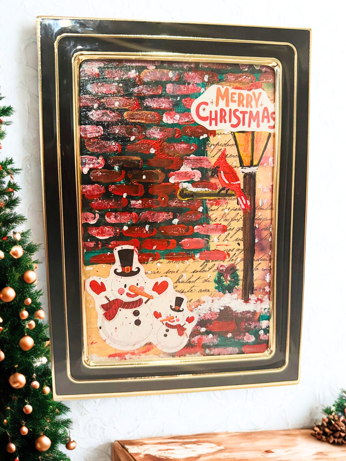 Framed Christmas-themed artwork with a snowman and festive text on a white wall.
