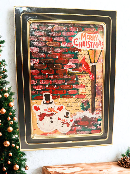 Framed Christmas-themed artwork with a snowman and festive text on a white wall.