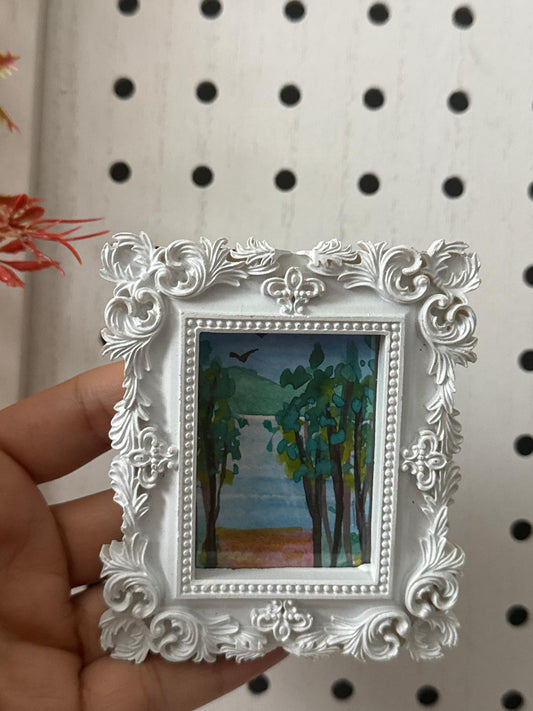 Decorative white frame with a small painting of a landscape held against a pegboard background.