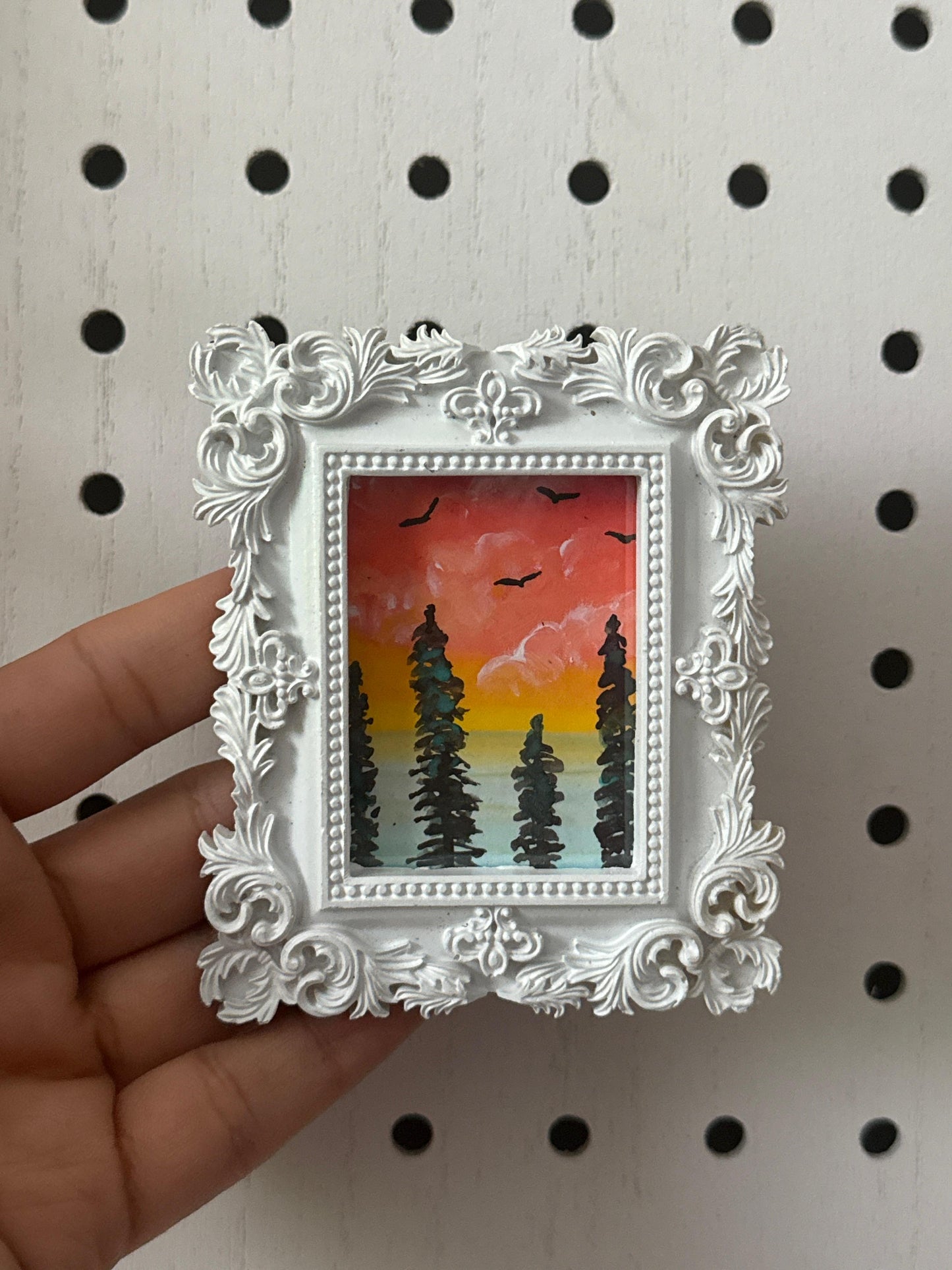 Small framed painting of a sunset with trees held against a pegboard background