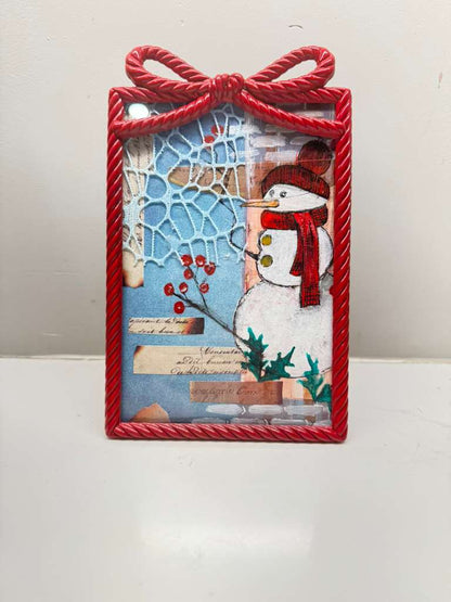 Handmade snowman Christmas artwork in a red bow frame with winter berries, snowflake texture, and vintage paper accents.