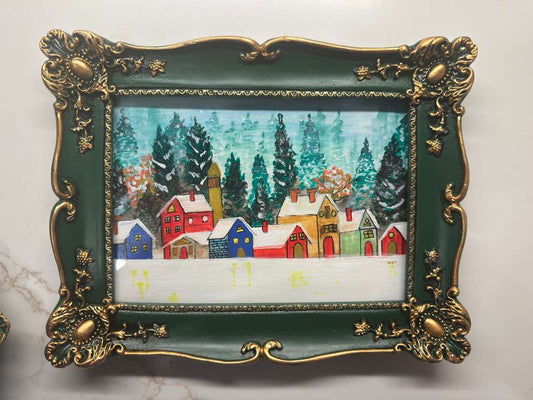Hand-painted winter village scene with colorful houses and pine trees in an ornate green and gold frame.