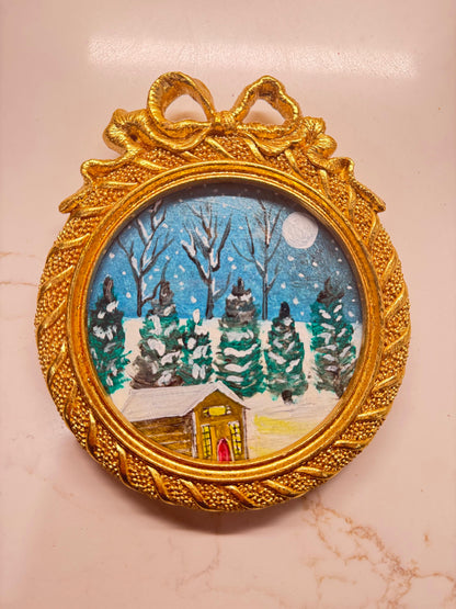 Handmade mini acrylic painting of a snowy winter cabin with pine trees and a full moon in a round gold ornament frame.