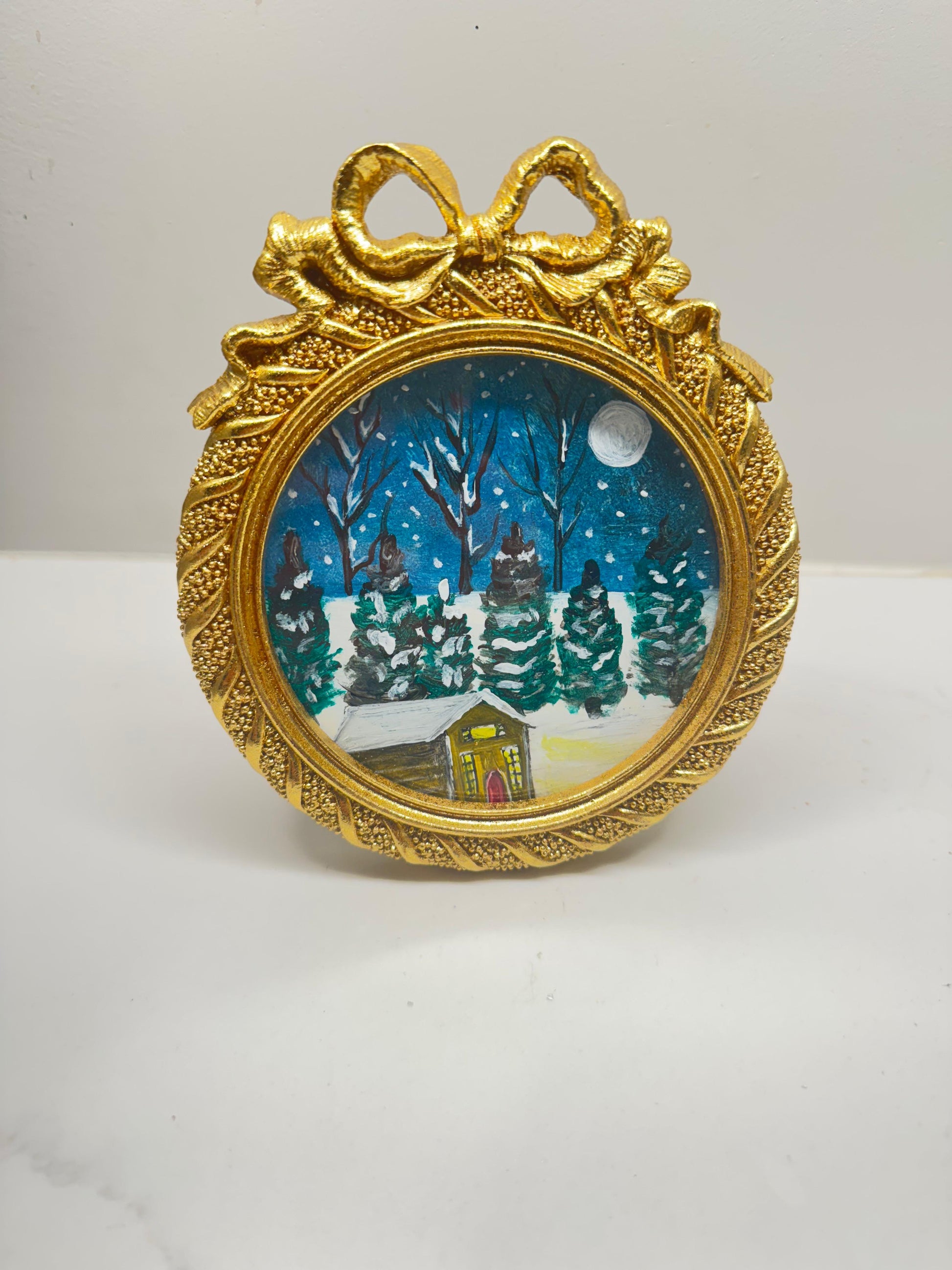 Handmade mini acrylic painting of a snowy winter cabin with pine trees and a full moon in a round gold ornament frame.