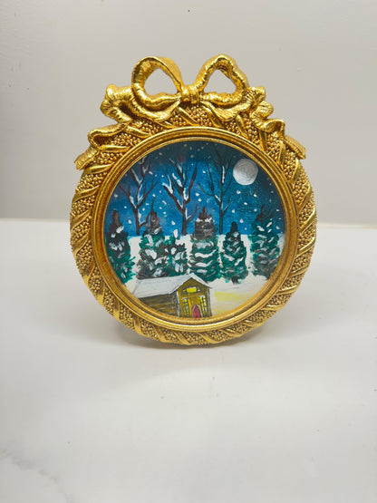 Handmade mini acrylic painting of a snowy winter cabin with pine trees and a full moon in a round gold ornament frame.