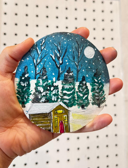 Handmade mini acrylic painting of a snowy winter cabin with pine trees and a full moon in a round gold ornament frame.