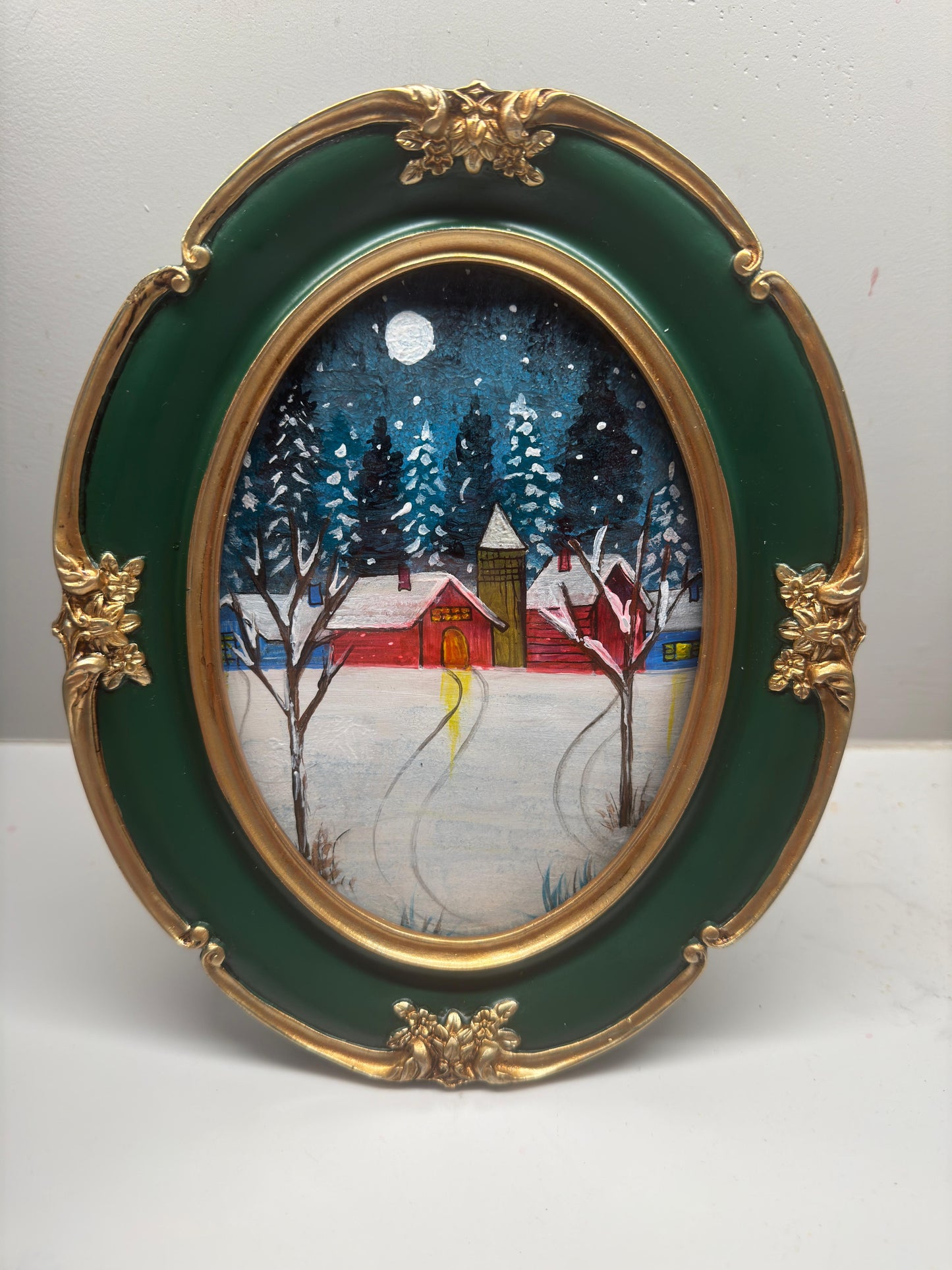 Mini acrylic painting of a red winter cabin with snow-covered trees and a full moon in a decorative green and gold oval frame.