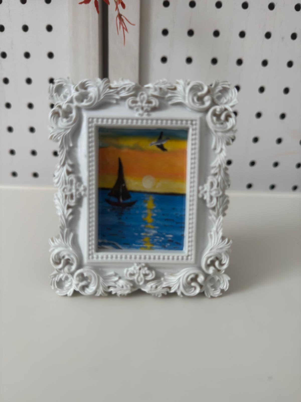 Decorative white frame with a small painting of a sailboat on a sunset background.