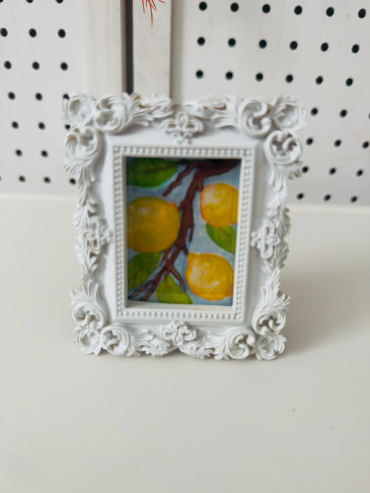 Decorative white frame with a painting of lemons on a white surface and black pegboard background
