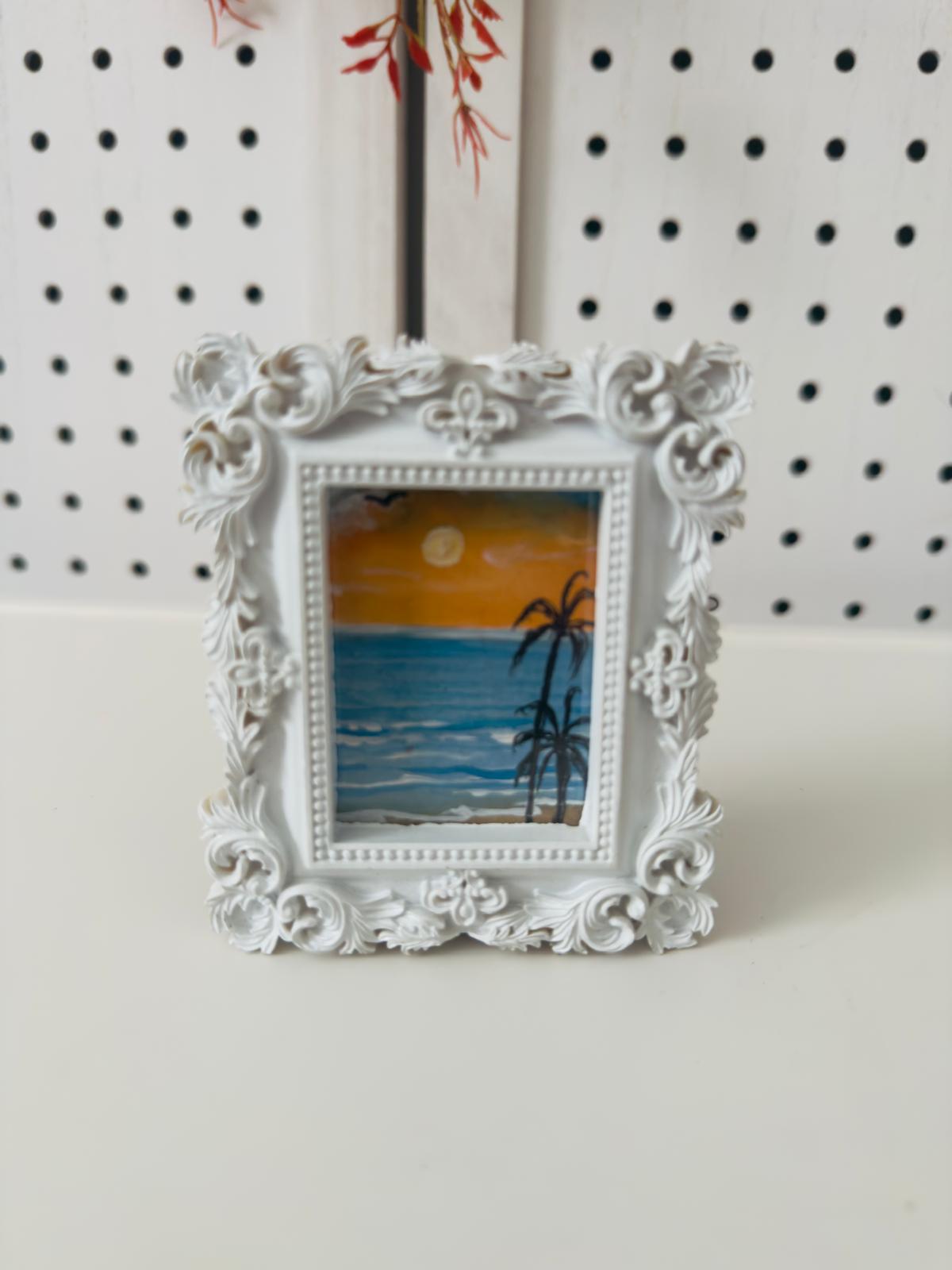 Decorative white frame with a small painting of a beach scene on a white surface.