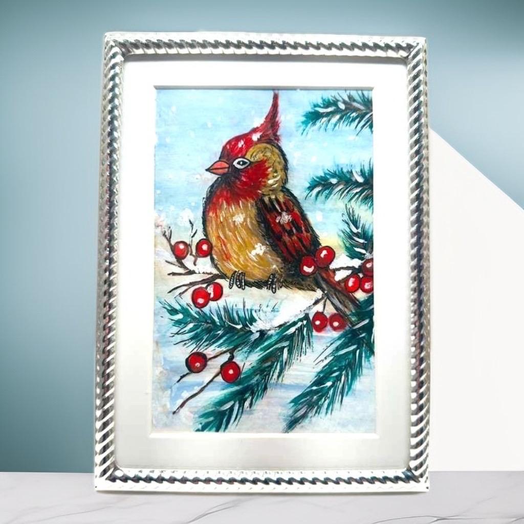 Holiday artwork of a winter cardinal bird with pine leaves and berries, set in a shiny silver picture frame.