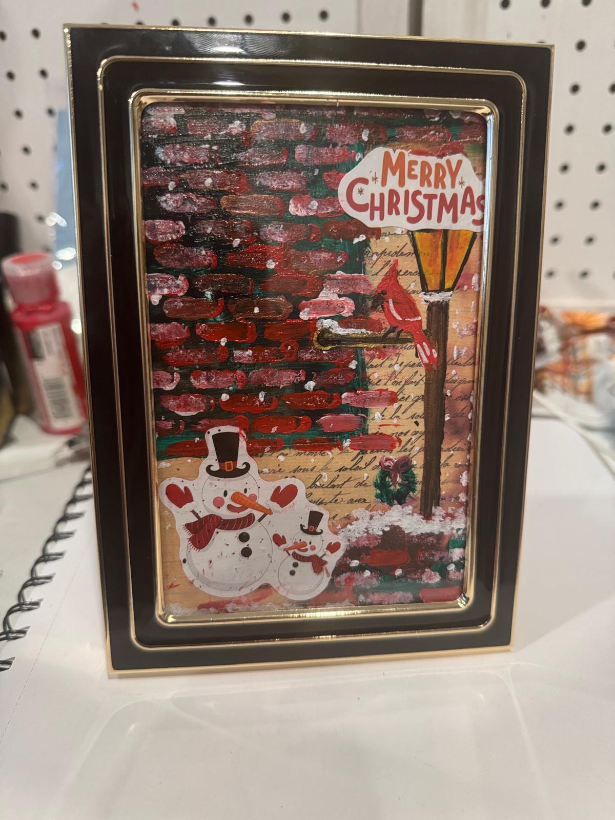 Framed Christmas card with a snowman and tree design on a white surface.