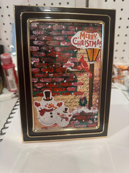 Framed Christmas card with a snowman and tree design on a white surface.