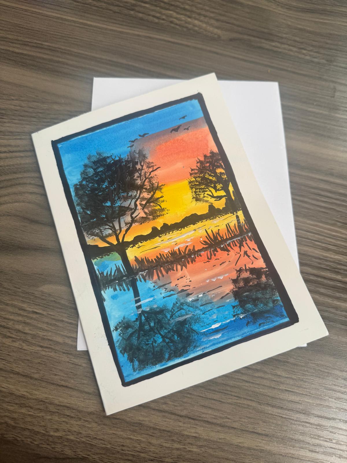 Handmade greeting card featuring colorful sunset over water with silhouetted trees .